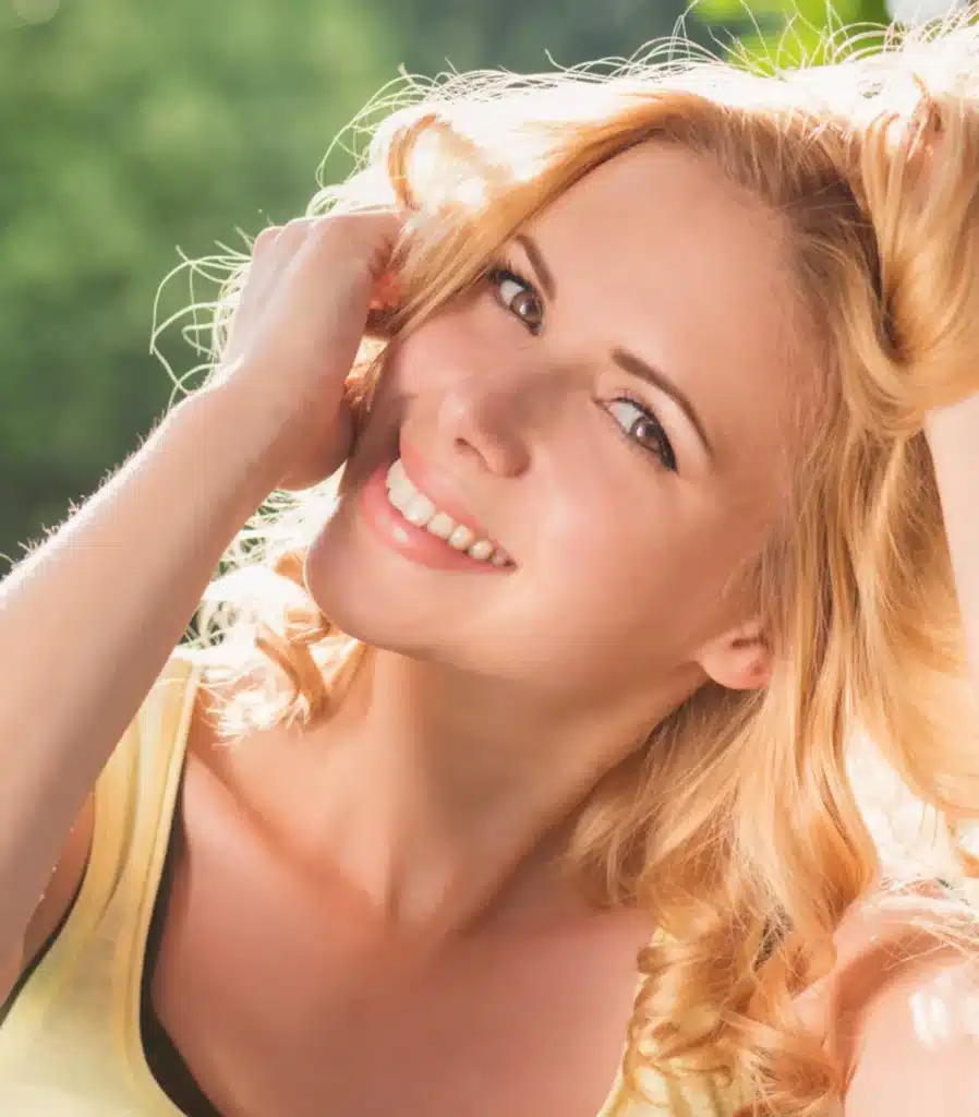 Mulher sorrindo em retrato de rosto, com cabelos loiros, em ambiente externo.